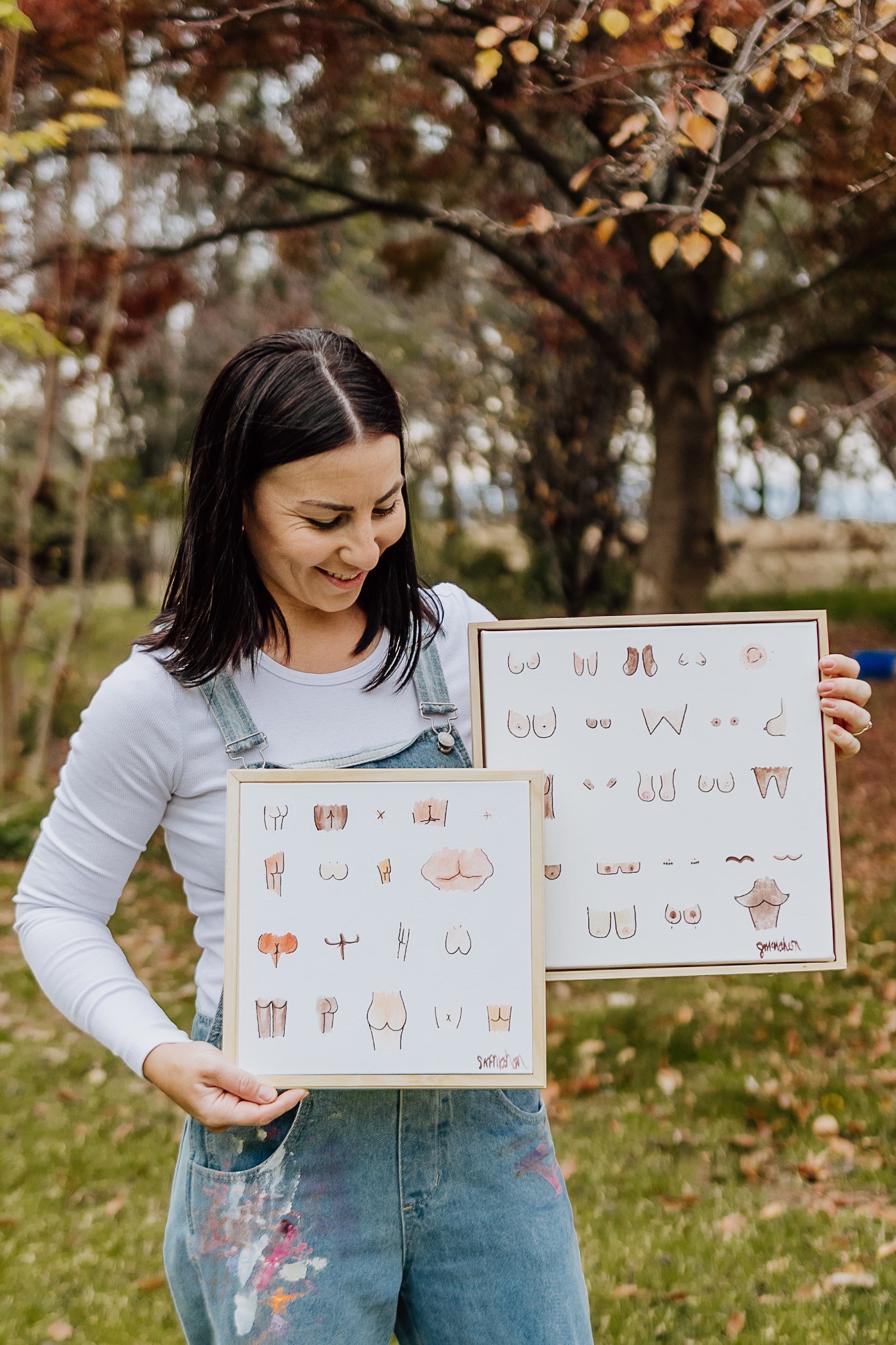 Woman holding two framed illustrations outdoors with trees and grass in the background